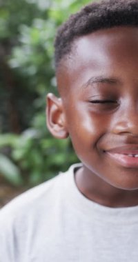 Dikey video: gülümseyen Afrikalı Amerikalı çocuk, yeşil yapraklı arka plan. Kısa siyah saçları, koyu kahverengi gözleri var ve sade gri bir gömlek giyiyor.