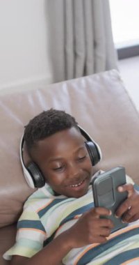 Dikey video: kulaklık takan, akıllı telefon tutan Afro-Amerikan çocuk. Kanepede uzanıyor, ekrana bakarken gülümsüyor, çizgili bir gömlek giyiyor.
