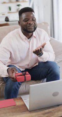 Dikey video: Afrikalı Amerikalı bir adam elinde hediye, dizüstü bilgisayarı önde. Kısa siyah saçları var, açık pembe gömlek giyiyor ve video çağrısında el hareketleriyle bir şeyler anlatıyor..