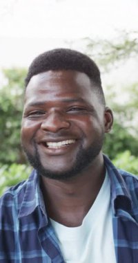 Dikey video: gündelik kıyafetler giyen, gülümseyen Afrikalı Amerikalı baba. Kısa siyah saçlı, sakallı ve beyaz tişörtünün üzerine mavi bir ekose gömlek giyiyor.