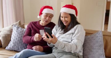 Noel zamanı, kıdemli Asyalı çift Noel Baba şapkası takıp akıllı telefon ve kredi kartıyla online alışveriş yapıyorlar. Çevrimiçi alışveriş, teknoloji, tatil, Noel, e-ticaret, festival