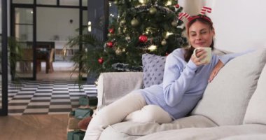 Kanepede dinlenen, şenlikli saç bandı takan bir kadın Noel ağacının yanında sıcak içkinin tadını çıkarıyor, fotokopi odası. tatil, rahatlama, kutlama, konfor, dekorasyon, ev