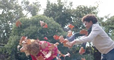 Mutlu Afro-Amerikan anne ve oğlu sıcak kıyafetler içinde bahçeye yapraklar fırlatıyor, ağır çekimde. Annelik, çocukluk, birliktelik, eğlence, sonbahar ve eğlence, değişmemiş.