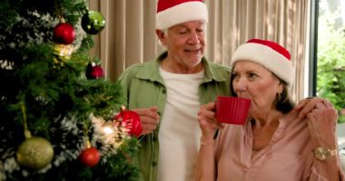 Noel Baba şapkalı yaşlı çift Noel ağacını süslüyor, evde neşeyle gülüyorlar. Tatil, şenlik, kutlama, kış, aile, birliktelik