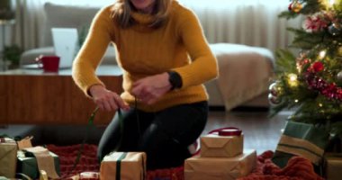 Noel Baba şapkalı gülümseyen kadın Noel ağacının yanında hediyeler paketliyor. Bayramlar, şenlikler, kutlamalar, hediyeler, dekorasyonlar, yaşam tarzı