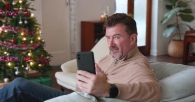 Evde süslü Noel ağacının yanında akıllı telefondan sohbet eden bir adam. Teknoloji, iletişim, kutlama, bayram, şenlik, dijital