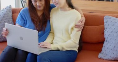 Gülümseyen Asyalı anne ve kızı evde dizüstü bilgisayar kullanarak kanepede oturuyorlar. Aile, bağ, teknoloji, birliktelik, internet, mutluluk