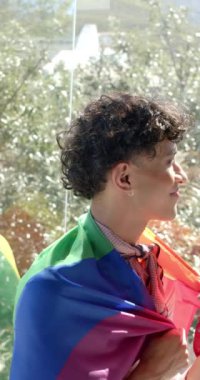 Dikey video: Gökkuşağı bayrağına sarılmış gülümseyen kişi pencere kenarında duruyor, kimliğini kucaklıyor. Gurur, LGBTQ, çeşitlilik, eşitlik, kapsayıcılık, neşe