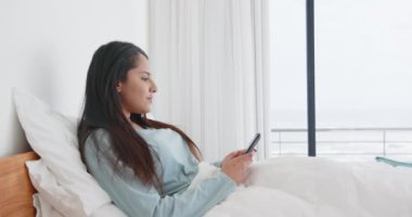 Yatakta dinlenen, Asyalı bir kadın akıllı telefon asistanı kullanıyor, evde okyanus manzarasının tadını çıkarıyor. Rahatlama, teknoloji, ses kontrolü, rahatlık, rahatlık, tatil