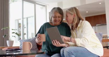 Tablet kullanarak, iki çok ırklı son sınıf kız arkadaş kanepede oturup evde kahve içiyorlar. Teknoloji, arkadaşlık, gevşeme, sohbet, sosyalleşme, rahatlama.