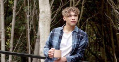 Ormanda kahvesini yudumlayan genç bir adam, günlük ekose gömlek giyiyor. Rahatlama, doğa, yaşam tarzı, vahşi doğa, boş zaman, macera.