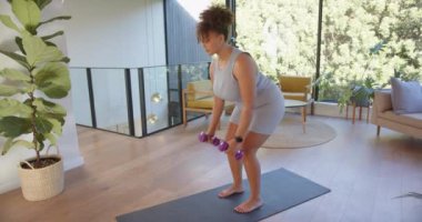 Dambıllarla egzersiz, ayrıca evde yoga minderiyle çalışan boyunda bir kadın. Spor, egzersiz, ev antrenmanı, dayanıklılık eğitimi, halter, sağlık.