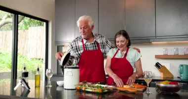 Son sınıf çifti mutfakta birlikte yemek pişiriyor, kırmızı önlük giyiyor ve evde sıcacık gülümsüyor. Son sınıflar, ev hayatı, mutluluk, yaşam tarzı, aşçılık, aile zamanı