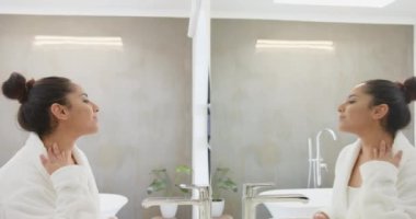 Aynaya bakıyor, bornozlu Asyalı kadın spa banyosunda boynuna dokunuyor, evde. Sağlık, rahatlama, kendine dikkat, güzellik, lüks, cilt bakımı.