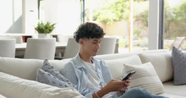 Kanepede oturmak, evde akıllı telefon kullanırken gülümseyen genç bir çocuk. Teknoloji, rahatlama, rahatlama, kapalı alan, gençlik, iletişim