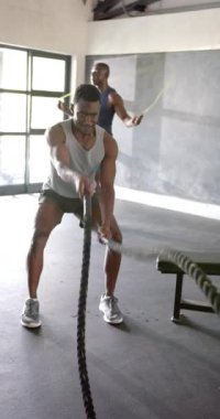 Dikey video: Erkekler dövüş halatlarıyla egzersiz yapıyor ve spor salonunda ip atlıyor, odaklanmış ve kararlı. Fitness, egzersiz, eğitim, güç, dayanıklılık, kararlılık