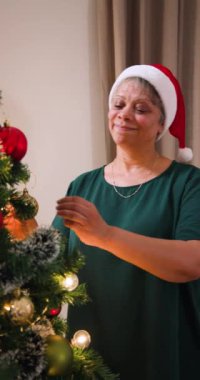 Dikey video: Yaşlı bir kadın Noel ağacını süslerle süslüyor, evinde sevinçle gülümsüyor. Tatil, şenlik, kutlama, gelenek, yaşlılık, mutluluk