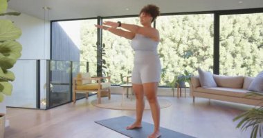 Yoga minderinde egzersiz, modern oturma odasında çömelmiş bir kadın. Fitness, egzersiz, ev çalışması, sağlık, vücut pozitifliği, aktif yaşam tarzı