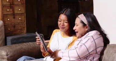 Çok ırklı genç bir kadın büyükannesine tablet gösteriyor. İkisi de evdeki rahat koltukta gülümsüyor. Teknoloji, aile, bağ, kuşak farkı, büyükanne ve büyükbaba, ev