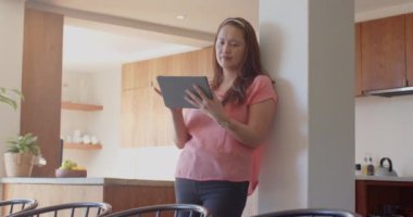 Tablet kullanıyor, kadın mutfakta dikiliyor, pencereden dikkatlice bakıyor. Teknoloji, yaşam tarzı, ev, tefekkür, modern, kapalı alan