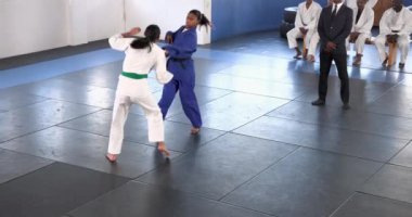 Judo antrenmanı yapan iki sporcu minder üzerinde antrenman yaparken diğerleri dikkatle izliyor. Dövüş sanatları, rekabet, eğitim, sportmenlik, takım çalışması, disiplin