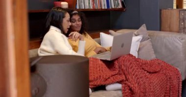 İki çok kültürlü bayan arkadaş kanepede dinleniyor, dizüstü bilgisayar izliyor, sıcak ev atmosferinin tadını çıkarıyorlar. Yaşam tarzı, eğlence, arkadaşlık, rahatlama, bağlanma, teknoloji