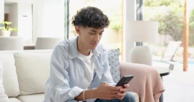 Kanepede oturan, akıllı telefon tutan ve evde gülümseyen genç bir çocuk. Teknoloji, rahatlama, rahatlama, yaşam tarzı, kapalı alan, konfor