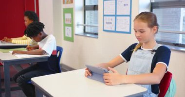 Okulda, masada tablet kullanan bir kız, arka planda okuyan sınıf arkadaşları. Eğitim, teknoloji, öğrenme, sınıf, çocuklar, öğrenciler