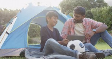 Açık havada kamp yapmak, Hintli baba ve oğul tablet kullanıyor ve futbol topu tutuyor. Aile, bağ, teknoloji, eğlence, eğlence, doğa.