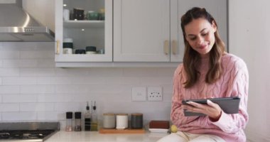 Evde, elinde tablet olan pembe kazaklı bir kadın, mutfakta düşünceli bir şekilde gülümsüyor, fotokopi çekiyor. Teknoloji, yaşam tarzı, dijital, iletişim, boş zaman, modern