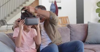 Hintli baba ve oğlu oturma odasındaki kanepede sanal gerçekliğin tadını çıkarıyorlar. Aile, bağ, teknoloji, eğlence, oyun, VR