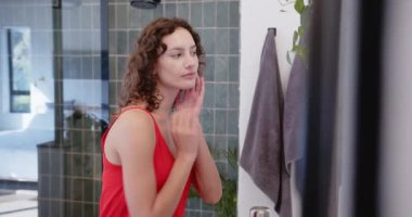 Cilt bakımı, kırmızı bluzlu kadın banyo aynasına bakıyor, gülümsüyor. Güzellik, kendine bakım, hijyen, kozmetik, bakım.