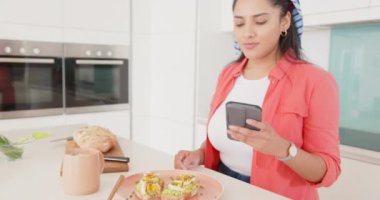 Mutfakta birden fazla iş yapmak, Asyalı bir kadının sandviç yemesi ve akıllı telefon kullanması. Yemek pişirme, teknoloji, meşgul, konfor, modern, yiyecek