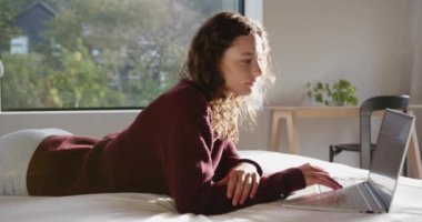 Yatakta uzanan, dizüstü bilgisayar kullanan ve rahat bir yatak odasında dinlenen bir kadın. Rahatlama, ev, yaşam tarzı, teknoloji, rahatlık, rahatlık