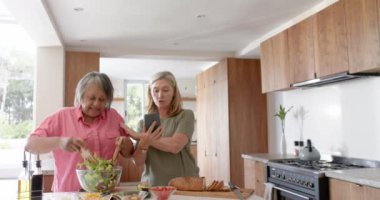 Salata hazırlamak, evde gülen ve akıllı telefon kullanan iki çok ırklı son sınıf kız arkadaş. yemek, arkadaşlık, yaşam tarzı, teknoloji, sağlıklı, yerli