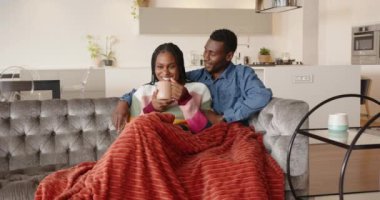 Kanepede dinlenen Afro-Amerikan çift evlerinde rahat bir battaniyenin altında kahvenin tadını çıkarıyor. Rahatlama, rahatlık, rahatlık, ev, yaşam tarzı.