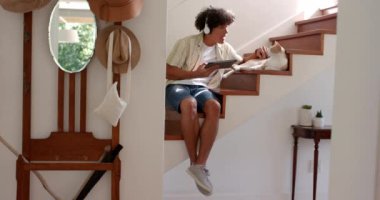 Merdivenlerde oturup, tablet kullanan ve rahat bir evde kediyle konuşan bir adam. Evcil hayvanlar, teknoloji, rahatlama, yerli, konfor, etkileşim