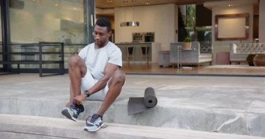 Merdivenlerde oturan Afro-Amerikan bir adam modern evin dışında yoga minderinin yanında ayakkabı bağlıyor. Spor, yaşam tarzı, egzersiz, açık hava, hazırlık, refah