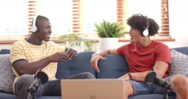 Müzik dinlerken, iki çok ırklı erkek arkadaş evde kulaklık ve dizüstü bilgisayarla rahatlıyorlar. teknoloji, yaşam tarzı, rahatlama, rahatlama, bağlanma, arkadaşlık