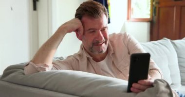 Son sınıf öğrencisi kanepede dinleniyor. Evde akıllı telefondan görüntülü konuşma yaparken gülümsüyor. Yaşlılar, teknoloji, iletişim, mutluluk, dijital, boş zaman