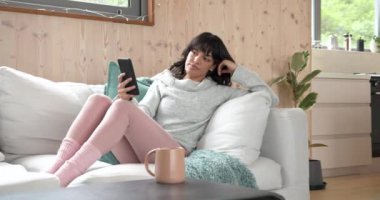 Evde dinlenirken, kadın rahat koltukta akıllı telefon kullanırken gülümsüyor. Teknoloji, rahatlama, rahatlık, konfor, yaşam tarzı, dijital