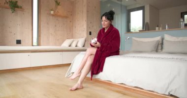 Kırmızı bornozlu kadın bacaklarına losyon sürüyor, rahat bir yatak odasında dinleniyor. Cilt bakımı, rahatlama, şımartma, güzellik, sağlık, yaşam tarzı.