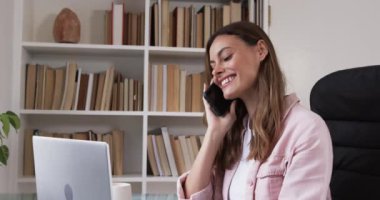 Evde bilgisayarla çalışırken akıllı telefondan konuşan gülümseyen kadın, kopyalama alanı. Uzaktan çalışma, iletişim, teknoloji, iş, ev ofisi, telekomünikasyon