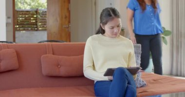 Kanepede oturan Asyalı anne ve kızı tablet kullanıyor ve birlikte gülümsüyor. Aile, bağ, teknoloji, rahatlama, ebeveynlik, mutluluk.