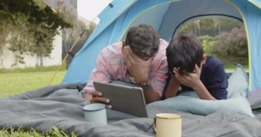 Çadırda kamp yaparken, Hintli baba ve oğul birlikte tablet izlerken gülüyorlar. Açık hava, bağ, aile, teknoloji, eğlence, doğa