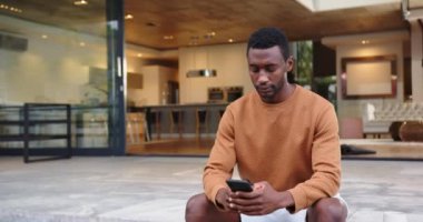 Modern evin dışında oturan Afro-Amerikan adam elinde akıllı bir telefonla düşünceli bir şekilde yüzünü çeviriyor. Teknoloji, yaşam tarzı, tefekkür, yalnızlık, rahatlama, açık hava.
