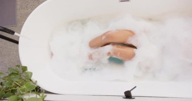 Küvette dinlenen, evinde köpük banyosunun keyfini çıkaran bir kadın. Rahatlama, kendine bakma, sağIık, kabarcıklar, sükunet, şımartma.