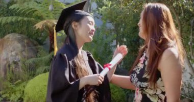 Mezun olan, kep ve cüppeli genç bir kadın Asyalı anneden diploma alıyor. Mezuniyet, başarı, tören, aile, kutlama, başarı