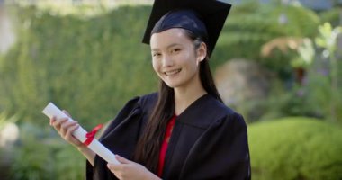 Mezun olan, şapkalı ve cüppeli genç Asyalı kız diplomasını tutuyor ve gülümsüyor. Mezuniyet, başarı, başarı, kutlama, mezuniyet, eğitim