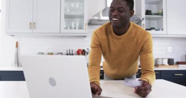 Gülümseyen adam, modern mutfakta kredi kartıyla online alışveriş yapıyor. E-ticaret, teknoloji, ödeme, yaşam tarzı, elverişli, işlem
